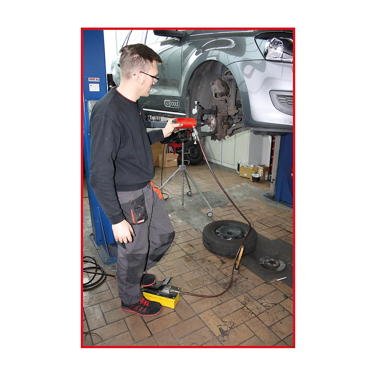 Ein Mechaniker repariert ein Auto in einer Werkstatt. Er benutzt ein Werkzeug, das an eine Pumpe angeschlossen ist. Ein Reifen liegt daneben.